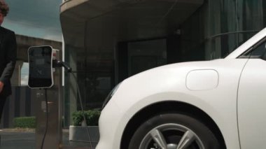 İlerici işadamı, şehir merkezinde dolaşmadan önce şarj cihazından elektrikli arabasına şarj prizi takıyor. Çevre dostu şarj edilebilir araba, sürdürülebilir ve temiz enerjiyle çalışıyor..