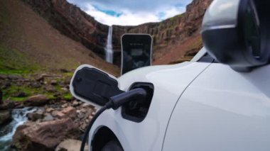 Elektrikli araba, doğal ortam, şelale ve akış arkaplanındaki şarj istasyonundan enerji yüklüyor. Yenilenebilir enerji kaynaklı EV otomobillerin enerji sürdürülebilirliği kavramı.