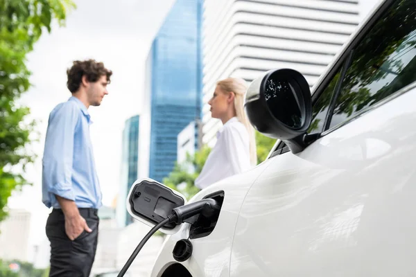 Focus parking-electric car connected to public charging station with blur progressive businesspeople holding coffee, residential building apartment and condo background for eco-friendly concept.