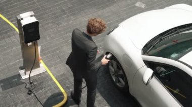 İlerici işadamı, şehir merkezinde dolaşmadan önce şarj cihazından elektrikli arabasına şarj prizi takıyor. Çevre dostu şarj edilebilir araba, sürdürülebilir ve temiz enerjiyle çalışıyor..