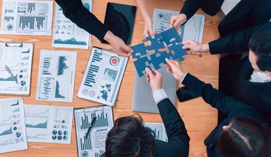 Top view businesspeople and colleagues in formal wear putting jigsaw puzzles together over meeting table with financial report papers in harmony office for team building concept.