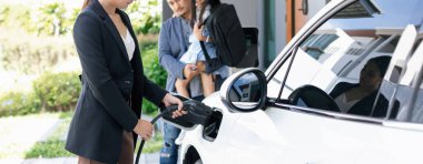 Progressive young parents and daughter with electric vehicle and home charging station. Green and clean energy from electric vehicles for healthy environment. Eco power from renewable source at home.