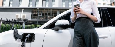 Closeup progressive suit-clad businesswoman with her electric vehicle recharge her car on public charging station in modern city with power cable plug and renewable energy-powered electric vehicle.