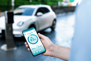 Closeup progressive businessman look at EV cars battery status application on smart phone screen at public parking charging station with power cable plug and renewable energy-powered electric vehicle
