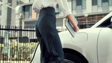 İlerici iş kadını şehir merkezinde dolaşmadan önce şarj cihazından elektrikli arabasına şarj prizi takıyor. Çevre dostu şarj edilebilir araba, sürdürülebilir ve temiz enerjiyle çalışıyor..