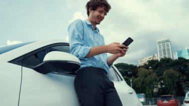 Takım elbiseli, hırslı bir işadamı elektrik kablosu ve yenilenebilir enerji enerjili bir elektrikli araçla şarj istasyonunda dururken elektrikli aracına yaslanıyor..