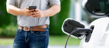 Progressive asian man install cable plug to his electric car with home charging station in the backyard. Concept use of electric vehicles in a progressive lifestyle contributes to clean environment.