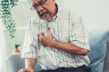 An agonizing senior man suffering from chest pain or heart attack alone in his living room. Serious health problem and feeling unwell concept for seniors.