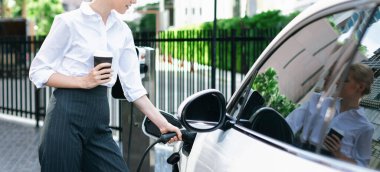 Elinde kahve olan kapanış işkadını kamu şarj istasyonunda elektrikli araca EV şarj cihazı takıyor. Şehirde ilerici yaşam tarzı için temiz ve yenilenebilir enerji kullanan çevre dostu bir araba..