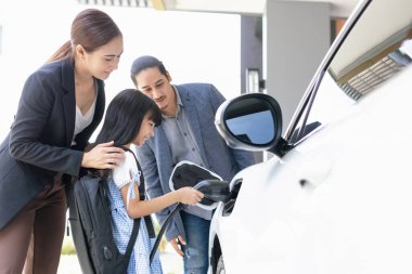 Progressive young parents and daughter with electric vehicle and home charging station. Green and clean energy from electric vehicles for healthy environment. Eco power from renewable source at home.