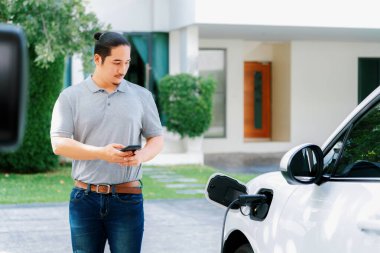 Progressive asian man install cable plug to his electric car with home charging station in the backyard. Concept use of electric vehicles in a progressive lifestyle contributes to clean environment.