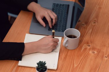 Closeup hand of secretary writing on notebook with laptop and coffee at her desk in harmony office. Businesswoman sitting on her workstation taking note in the meeting for financial strategy planning.