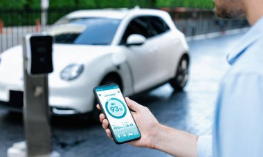 Closeup progressive businessman look at EV cars battery status application on smart phone screen at public parking charging station with power cable plug and renewable energy-powered electric vehicle