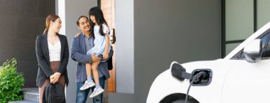 Progressive young parents and daughter with electric vehicle and home charging station. Green and clean energy from electric vehicles for healthy environment. Eco power from renewable source at home.