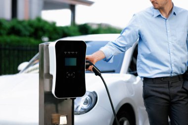 Closeup progressive man holding EV charger plug from public charging station for electric vehicle with background of residential building as concept eco-friendly sustainability energy car concept.