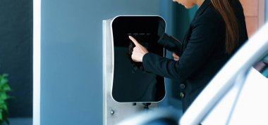 İlerici kadın elektrikli arabasının fişini şarj istasyonuna takıyor. İlerici bir yaşam tarzında elektrikli araç kullanımı kavramı temiz çevreye katkıda bulunur.