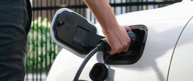 Closeup progressive man holding EV charger plug from public charging station for electric vehicle with background of residential building as concept eco-friendly sustainability energy car concept.