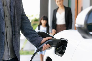 Elektrikli arabaya odaklan, elektrikli araca şarj cihazı yerleştir. Arka planda bulanık aile ile alternatif yeşil enerji teknolojisi kavramını geliştir..