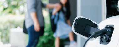 Arka planda bulanık baba ve kızının yürüdüğü şarj istasyonunda elektrikli araba şarj etmeye odaklan. Sağlıklı çevre yaşam tarzı konsepti için yenilikçi yeşil ve temiz enerji aracı.