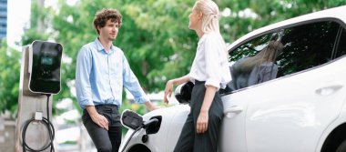 Progressive businessman and businesswoman with electric car parking and connected to public charging station before driving around city center. Eco friendly rechargeable car powered by clean energy.