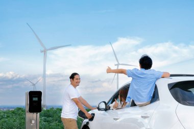 İlerici mutlu aile kavramı elektrikli araçla rüzgar çiftliğinde eğleniyor. Rüzgar türbini jeneratöründen gelen temiz yenilenebilir enerji ile şarj edilen elektrikli araç..