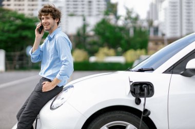 Progressive businessman talking on the phone, leaning on electric car recharging with public EV charging station, apartment condo residential building on the background as green city lifestyle.