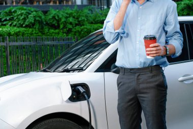 Progressive eco-friendly concept of parking EV car at public electric-powered charging station in city with blur background of businessman leaning on recharging-electric vehicle with coffee.
