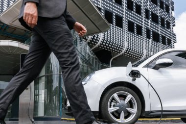 Closeup progressive businessman with electric car recharging at public charging station with background of city residential building. Eco friendly car powered by alternative clean energy.