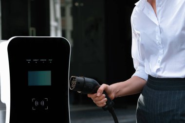 Focus EV charger plug and electric car at public charging station with blur progressive businesswoman holding charger and apartment condo building in background. Eco friendly electric vehicle concept.