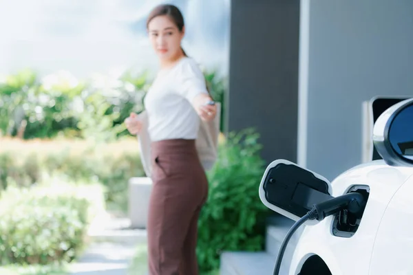 Focus EV charger plugged into EV car at home charging station with blurred background of progressive woman walking in background. Elective vehicle powered by clean energy for eco-friendly concept.