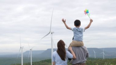 İlerici mutlu aile kavramı rüzgâr türbini çiftliğinde eğleniyor. Rüzgar türbini jeneratörüyle gelen elektrik jeneratörü kırsal bölgede tepe ve ufukta dağ var..