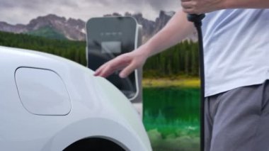 İlerici adam şarj noktasında elektrikli araba yüklüyor, daha temiz bir çevre için temiz ve yenilenebilir enerjiyle çalışan bir EV ile doğada ve dağlarda seyahat ediyor..