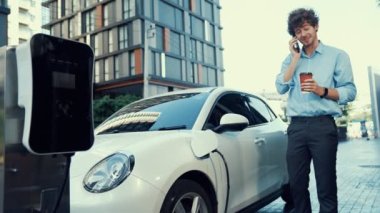 Takım elbiseli, hırslı bir işadamı elektrik kablosu ve yenilenebilir enerji enerjili bir elektrikli araçla şarj istasyonunda dururken elektrikli aracına yaslanıyor..