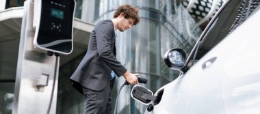 Below view closeup image of progressive black suit businessman recharge battery of his electric vehicle from public charging station. Renewable and alternative energy powered car concept.