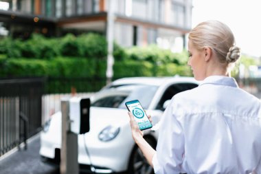 Suit-clad progressive businesswoman look at EV cars battery status from her phone while standing on a charging station with a power cable plug and a renewable energy-powered electric vehicle.