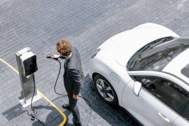 Aerial view of progressive businessman in black formal suit with his electric vehicle recharging battery at public car park charging station as vehicle powered by sustainable energy concept.