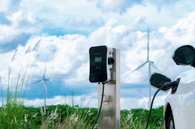 Progressive combination of wind turbine and EV car, future energy infrastructure. Electric vehicle being charged at charging station powered by renewable energy from wind turbine in the countryside.