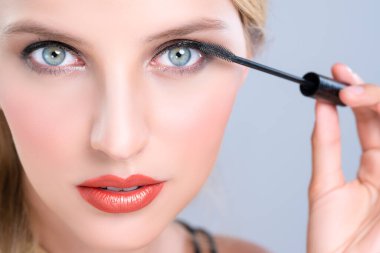 Closeup woman with blond hair putting alluring black mascara with brush in hand on long thick eyelash. Perfect fashionable cosmetic clean facial skin with beautiful eye young woman in high resolution.