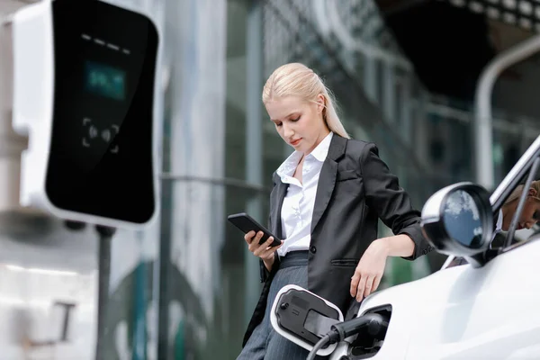 Businesswoman Wearing Black Suit Using Smartphone Leaning Electric Car Recharge — Stockfoto