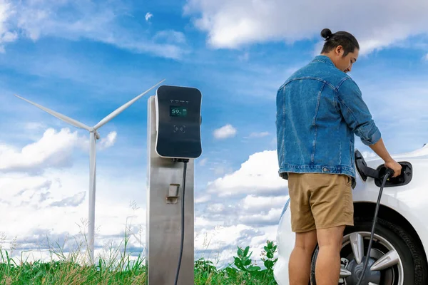 电动汽车是一个进步的人 他的电动汽车 电动汽车用风力涡轮机作为未来可持续能源的概念 从绿地的充电站向能源充电 配备发电机的电动车 — 图库照片