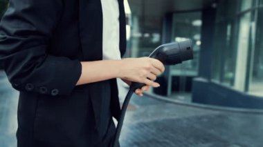 İlerici iş kadını şehir merkezinde dolaşmadan önce şarj cihazından elektrikli arabasına şarj prizi takıyor. Çevre dostu şarj edilebilir araba, sürdürülebilir ve temiz enerjiyle çalışıyor..