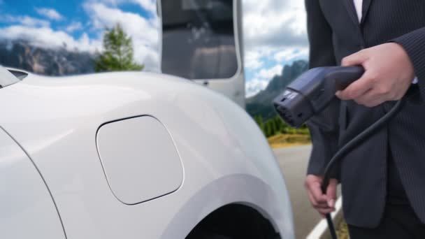 ビジネススーツの進歩的な男は山と田舎の国の充電ステーションから彼の電気自動車を充電します 道路旅行 再生可能エネルギーを搭載したEv車での冒険 — ストック動画