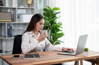 Modern ofis masasındaki Asyalı hevesli genç iş kadını bir fincan kahveyle çalışmak için dizüstü bilgisayar kullanıyor. Çalışkan ve çekici ofis hanımı ofisinde bilgisayar defteri üzerinde çalışıyor..
