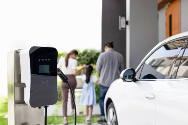 Focus closeup electric vehicle recharging battery from home electric charging station with blurred family in background. Renewable clean energy car for progressive eco awareness lifestyle concept.