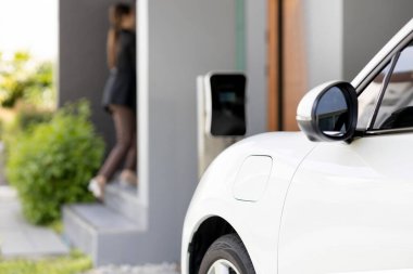 Focus image of electric vehicle recharging battery at home charging station with blurred woman walking in the background. Progressive concept of green energy technology applied in daily lifestyle.