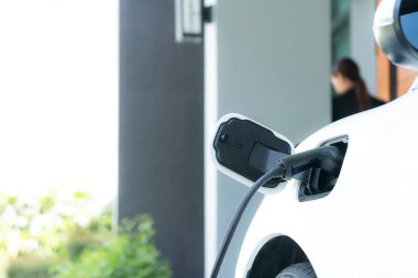 Focus image of electric vehicle recharging battery at home charging station with blurred woman walking in the background. Progressive concept of green energy technology applied in daily lifestyle.