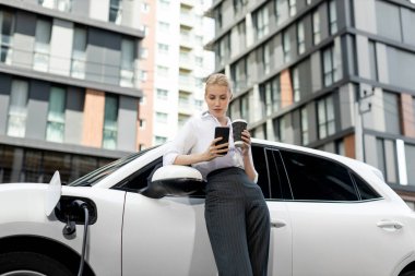 Focus businessman using phone, leaning on electric vehicle, holding coffee with blurred city residential condo buildings in background as progressive lifestyle by renewable and sustainable EV car.