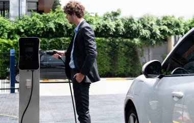 Modern şehir yerleşim bölgesindeki kamu şarj istasyonunda elektrikli araba şarj eden yenilikçi bir işadamı. Çevre dostu şarj edilebilir araba alternatif temiz enerjiyle çalışıyor..