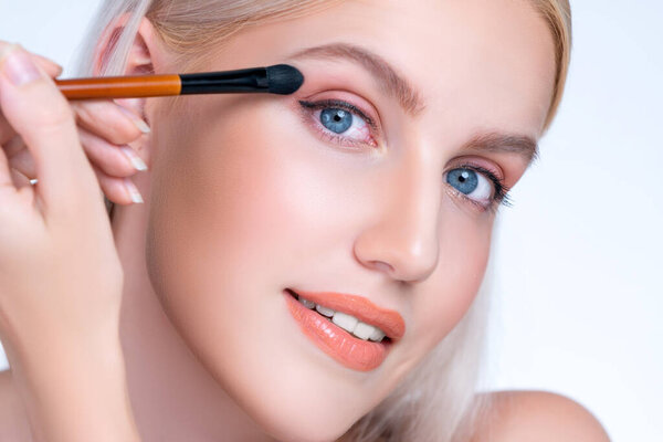 Крупный план красивая представительная девушка с безупречным нанесением тени для глаз макияж с кистью подводки для глаз. Процесс косметической живописи лица на красивой молодой женщине с идеальной чистой кожей на изолированном фоне.