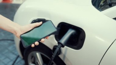 Taklit uzay için şarj istasyonuna bağlı elektrikli aracın yeşil ekran enerji durumunu gösteren bir model telefon. Temiz çevre için ilerici bir kavram. EV, yeşil yenilenebilir enerjiden güç alıyor.